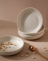 A white stoneware pasta plate with a matte finish. The plate is empty, and there are scattered chickpeas on the surface with a wooden spoon to the side.