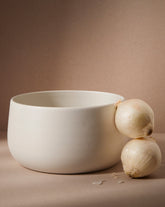 A large white stoneware serving bowl with a minimalist design, accompanied by two onions, presented against a neutral background.
