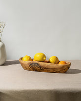 An oval-shaped, rustic bowl made of olivewood, displaying natural wood patterns, filled with lemons, placed on a table with a neutral cloth.