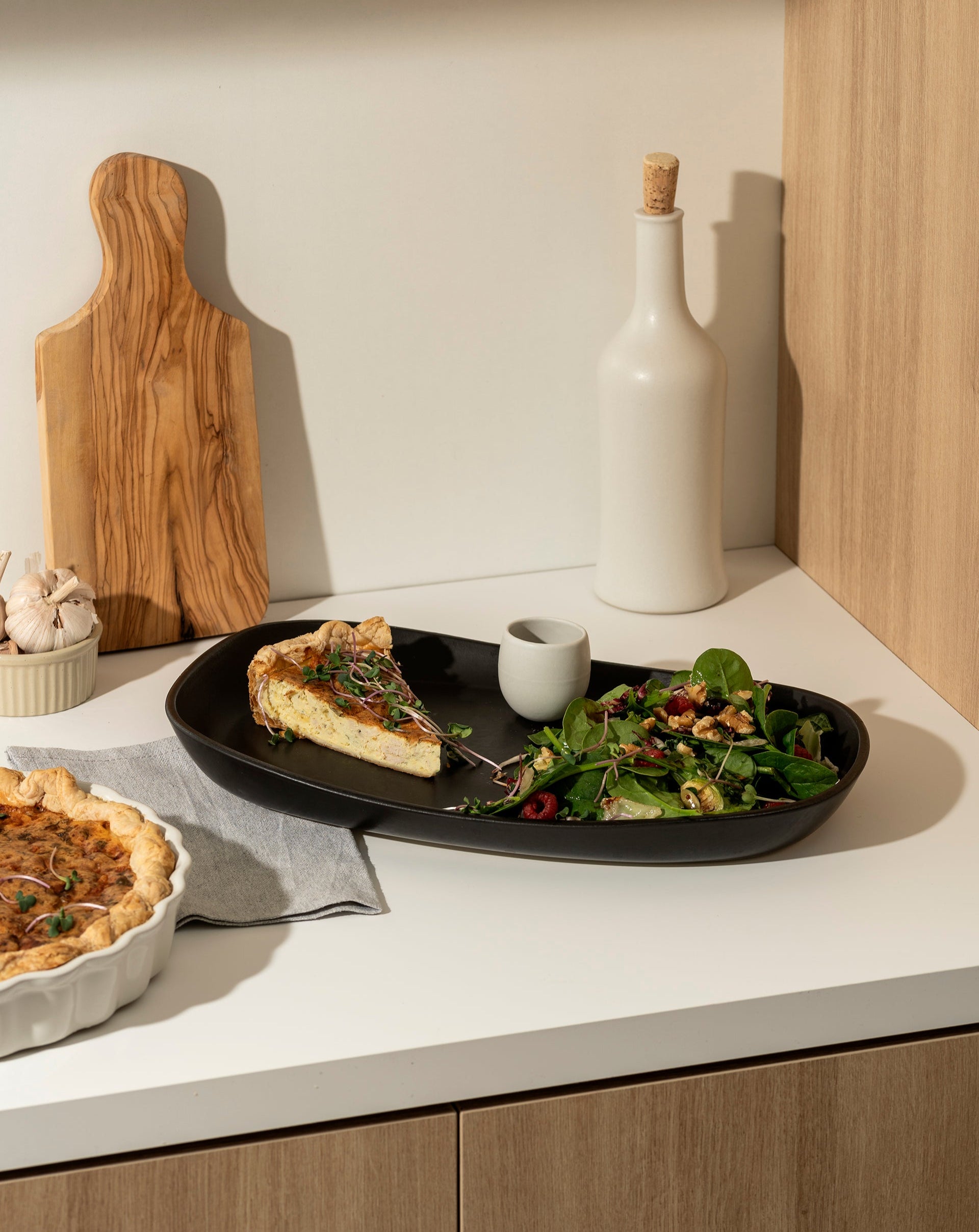 A kitchen counter with a black plate holding a slice of quiche, a green salad, and a small cup. Nearby are a white bottle with a cork, a wooden cutting board, garlic, and a whole quiche in a white dish