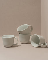 A set of four white stoneware coffee mugs, three stacked behind one in front, on a beige background.
