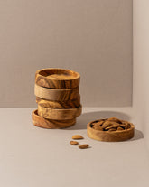 Five stacked olive wood bowls beside a separate bowl filled with walnuts, with a few scattered walnuts on the brown surface.