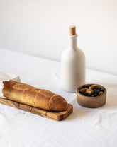 An olive oil bottle and a loaf of bread on a wooden board, with a small wooden bowl of olives beside it, all placed on a white surface.