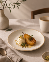 A beautifully styled dining table setting featuring a ceramic plate with a golden pear and a scoop of ice cream topped with crushed pistachios, alongside a vase, spoon, and cup on a linen tablecloth.