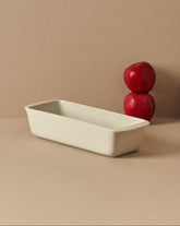 A white enameled stoneware cake and bread pan on a beige background, with two red apples placed on top of it to the side.