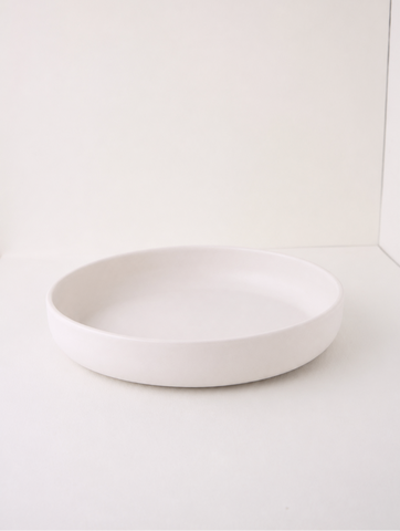 A minimalist, shallow round ceramic plate in matte white, photographed against a clean white background.