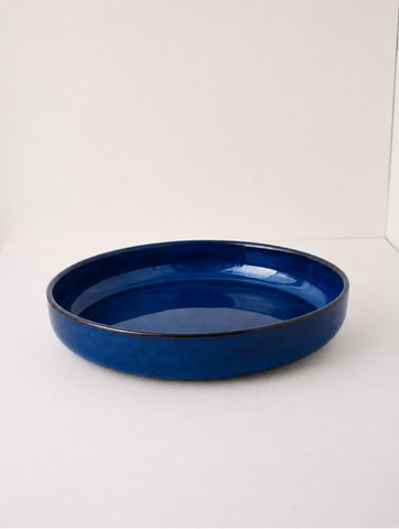 A glossy cobalt blue ceramic shallow bowl or dish, photographed from a side angle against a plain white studio background.