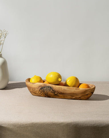 An oval-shaped, rustic bowl made of olivewood, displaying natural wood patterns, filled with lemons, placed on a table with a neutral cloth.