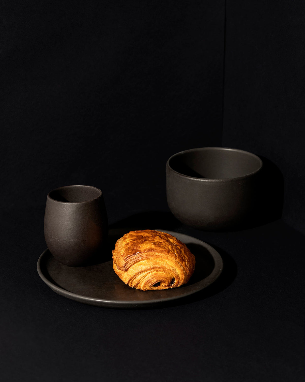 A minimalist white dinnerware set displayed against a neutral background, including a flat salad plate with a dessert, a dinner bowl, and a cup.