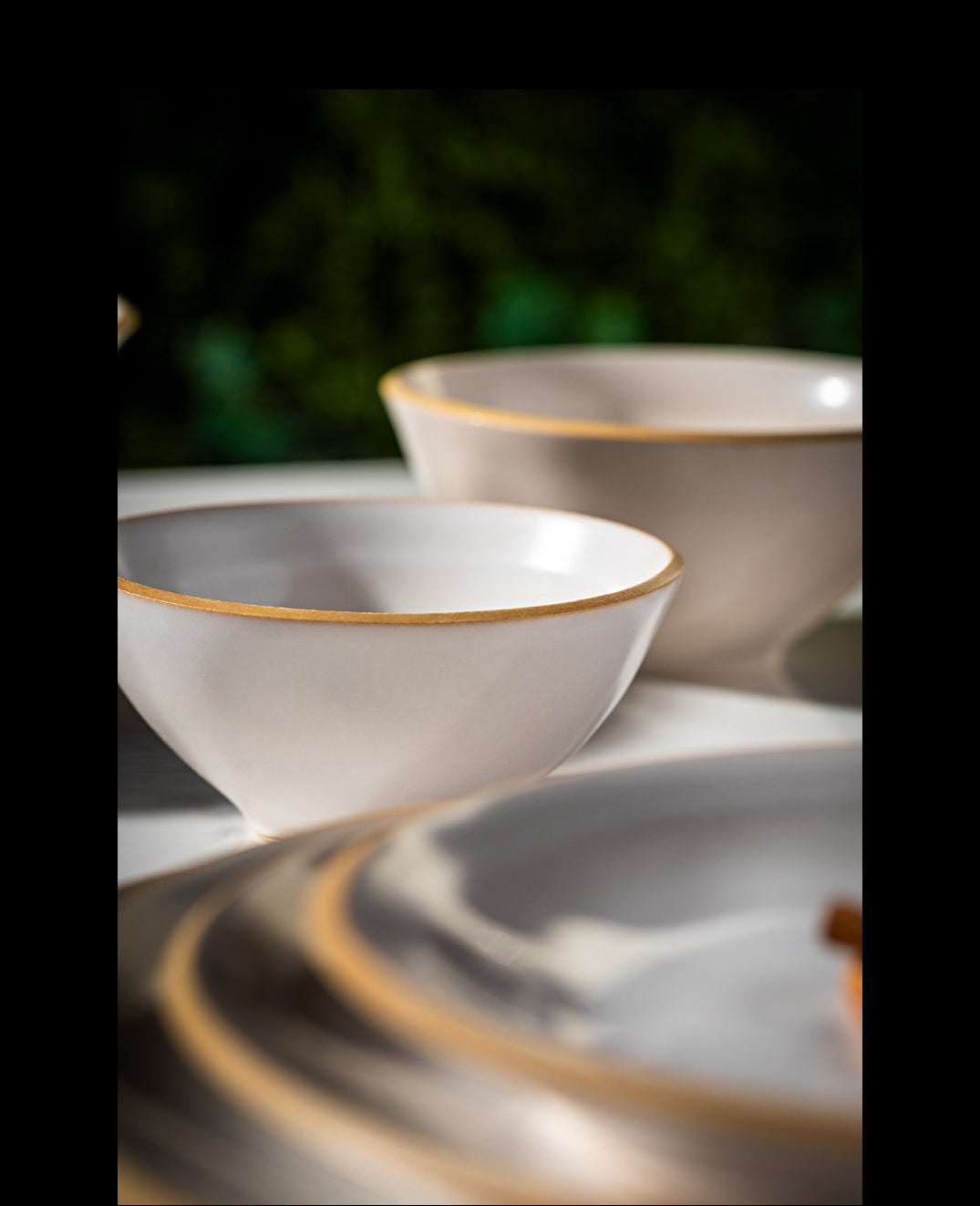 White ceramic bowls with a thin gold rim, shown in a close-up shallow depth-of-field shot on a table with matching plates and a blurred green background.