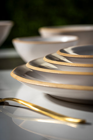 A close-up, artistic shot of a stack of white ceramic plates with a thin gold rim, alongside a matching gold spoon, set against a dark, blurred background.