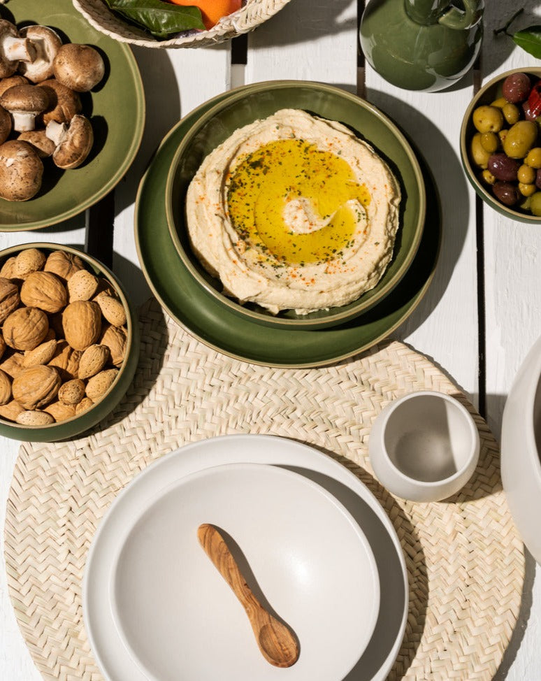 A beige palm fiber placemat with two stacked plates and a wooden spoon resting on top. Surrounding the setup are additional bowls filled with various food items, creating a warm, natural tablescape.